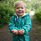 A blonde toddler smiles, holding out his hands, wearing a green 3 in 1 Waterproof Scampsuit Green with a white lightning bolt pattern.